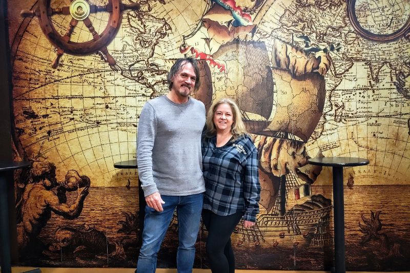 Brewery owners Rick and Cynthia Lee pose in front of an interior wall adorned with pirate ships inside DarkHeart Brewing